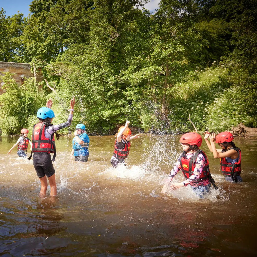 Six children wearing helmets and life jackets play and splash in a river.