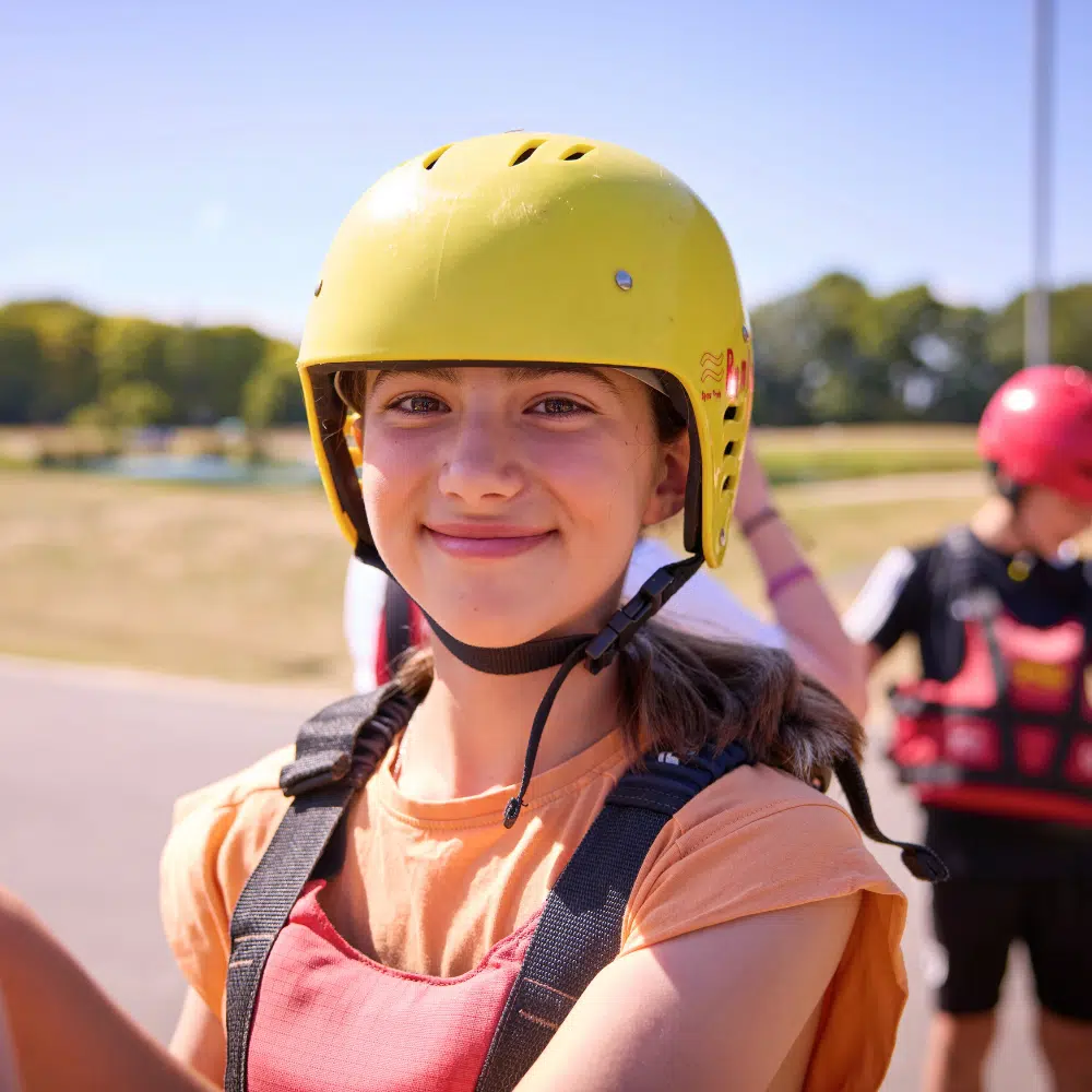 A girl wearing a yellow helmet and a life jacket smiles at the camera.