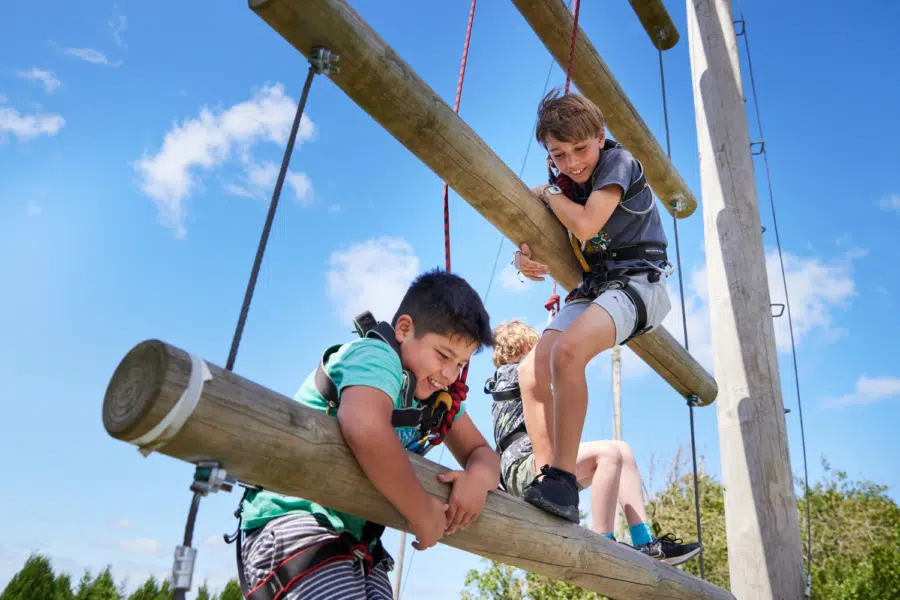 Two children in harnesses navigate a high ropes course, gripping wooden beams suspended in the air.
