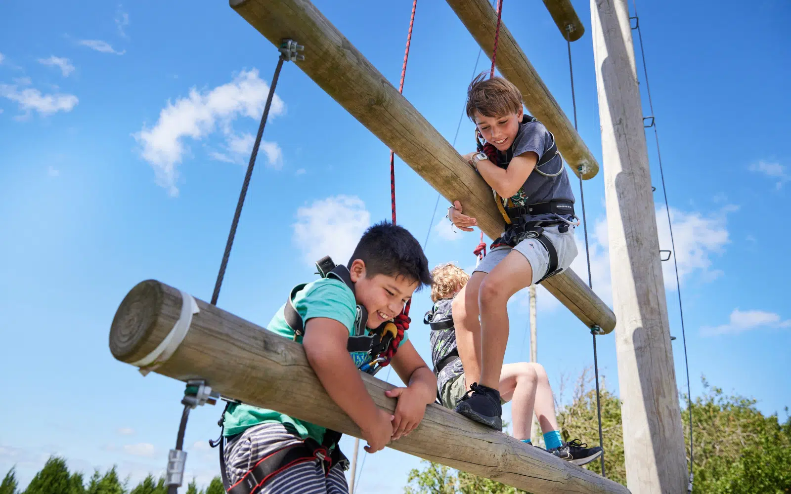 Two children in harnesses navigate a high ropes course, gripping wooden beams suspended in the air.